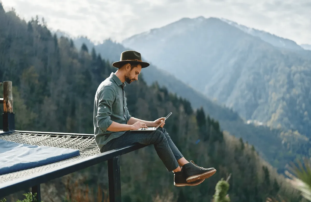 Mann arbeitet mit Laptop auf einem Steg vor einem Bergpanorama